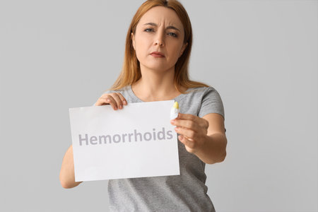 Mature woman holding paper with word HEMORRHOIDS and rectal suppository on light backgroundの写真素材