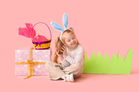 Cute little girl in bunny ears with chocolate Easter egg, gifts and paper grass on pink background