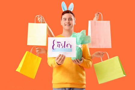 Young man in bunny ears with gift egg and shopping bags on orange background. Easter Sale