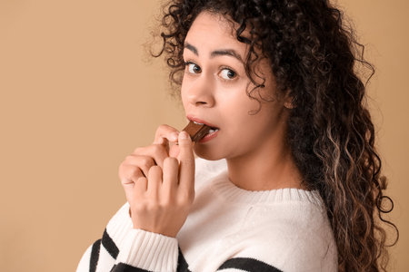 Beautiful young African-American woman eating piece of sweet chocolate on brown backgroundの写真素材