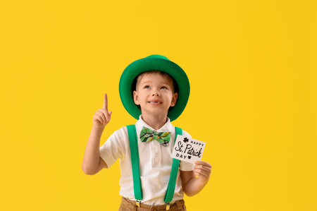 Cute little boy with leprechaun's hat and greeting card pointing at something on yellow background. St. Patrick's Day celebrationの写真素材