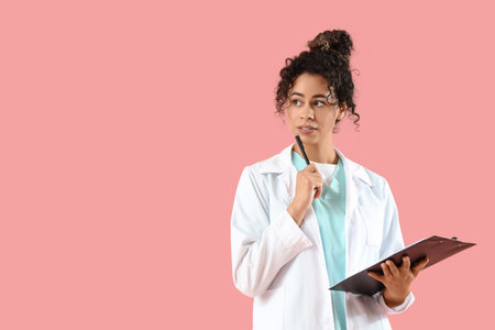 Thoughtful female African-American medical intern with clipboard on pink backgroundの写真素材