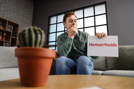 Young man holding paper with the word HEMORRHOIDS on sofa at homeの写真素材