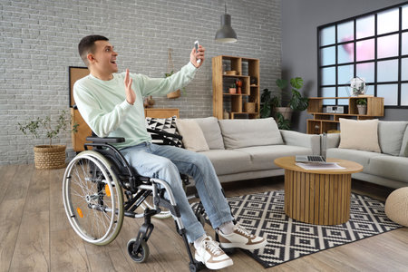 Happy young man in wheelchair with mobile phone taking selfie at homeの写真素材