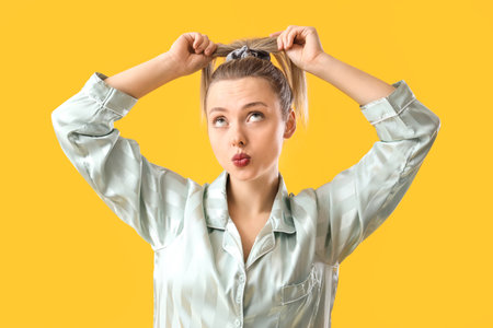 Young blonde woman in pajamas doing ponytail with scrunchy on yellow backgroundの写真素材