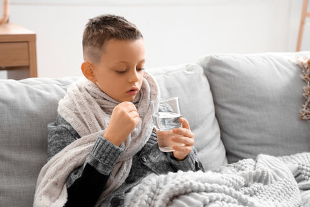 Ill little boy with glass of water taking pill on sofa at homeの写真素材