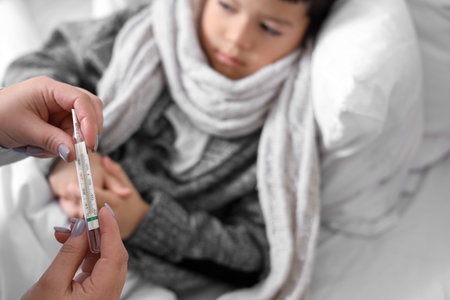Mother with thermometer and her ill little son in bedroom, closeup