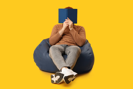 Man sitting on beanbag and covering his face with book against yellow backgroundの写真素材