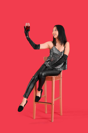 Beautiful Asian woman with glass of wine sitting on red backgroundの写真素材
