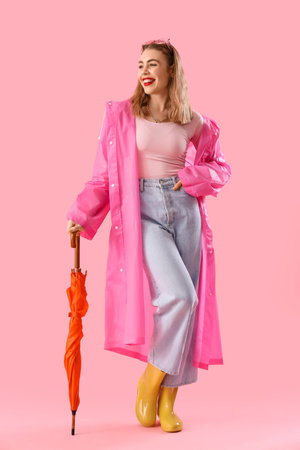 Beautiful stylish woman in raincoat with umbrella on pink backgroundの写真素材