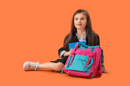 Cute little schoolgirl with book and backpack sitting on orange backgroundの写真素材