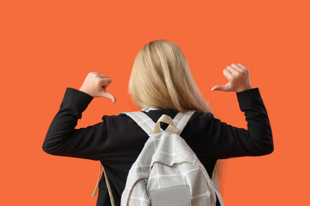 Little schoolgirl pointing at backpack on orange background, back viewの写真素材