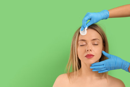 Cosmetologist with cotton pad removing makeup from young woman's face on green backgroundの写真素材