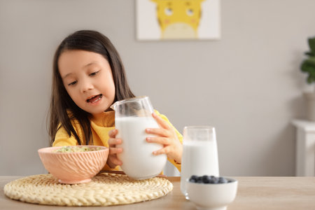 Cute little Asian girl pouring milk into bowl of cereal rings at table in kitchenの写真素材