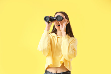 Young woman looking through binoculars on yellow backgroundの写真素材