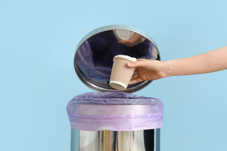Female hand throwing used paper cup into trash bin near blue wall