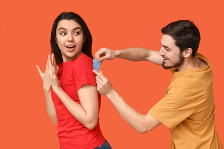 Young man sticking paper fish on his friend's back against orange background. April fool's day celebrationの写真素材
