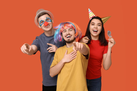 Young friends in funny disguise with paper fish pointing at viewer on orange background. April...