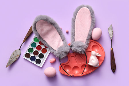 Bunny ears with Easter eggs and painter supplies on lilac background