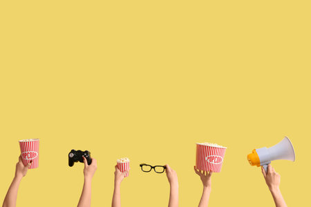 Hands holding buckets with popcorn, 3D glasses, game pad and megaphone on yellow backgroundの写真素材