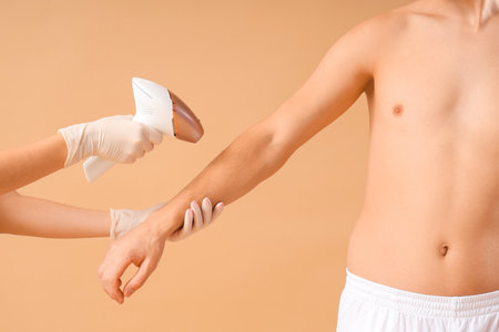 Cosmetologist using photoepilator for removal of hair from man's body on beige backgroundの写真素材