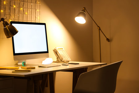 Blank computer monitor with stationery and glowing lamps in office at nightの写真素材