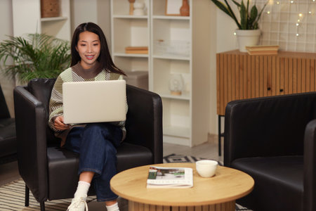 Young Asian woman using laptop on black armchair at home in eveningの写真素材