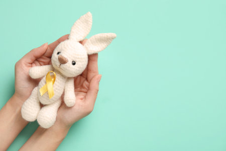 Female hands with toy bunny and golden awareness ribbon on turquoise background. International Childhood Cancer Dayの写真素材