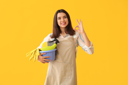 Young woman with cleaning supplies showing OK on yellow backgroundの写真素材
