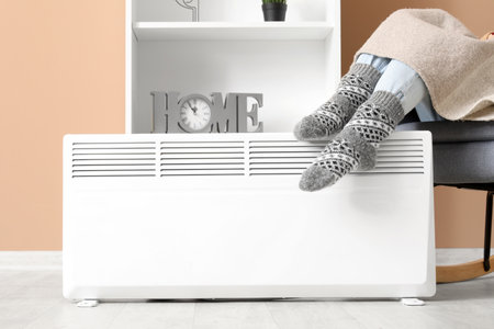 Young woman in socks with warm plaid near electric heater at home. Concept of heating seasonの写真素材