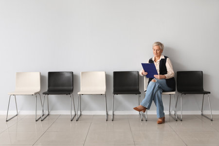 Mature female applicant with clipboard waiting for job interview near light wallの写真素材