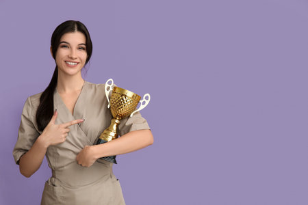 Young cosmetologist pointing at gold cup on lilac backgroundの写真素材