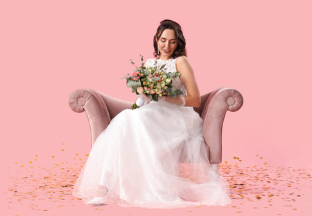 Beautiful bride with wedding bouquet in armchair on pink backgroundの写真素材