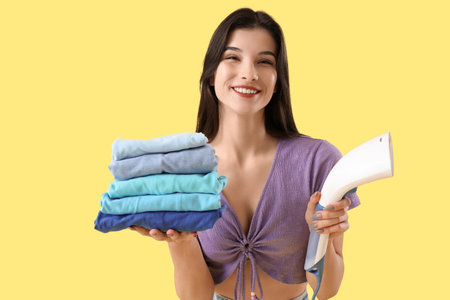 Pretty young woman with modern garment steamer and stack of clean clothes on yellowの写真素材