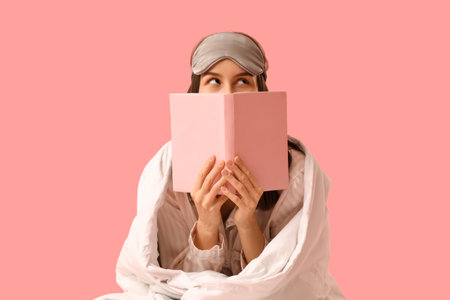 Pretty young woman with blanket, sleeping mask and reading book on pink backgroundの写真素材