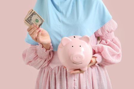 Beautiful young Muslim woman with piggy bank and dollars on pink background, closeupの写真素材