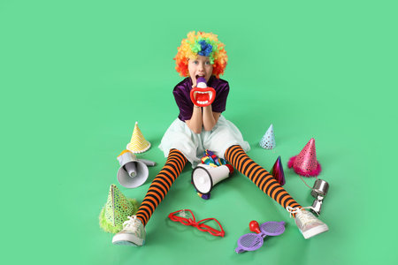 Funny girl in clown costume with party hats and megaphones on green background. April Fool's Day...