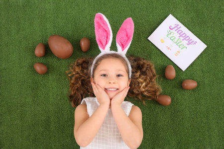 Cute little girl in bunny ears with chocolate Easter eggs and card on green grass, top view