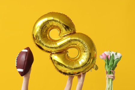 Female hands with foil balloon in shape of figure 8, rugby ball and tulip flowers for International Women's Day on yellow backgroundの写真素材