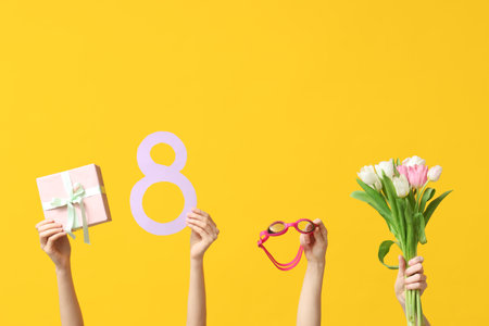 Female hands with paper figure 8, goggles, gift box and tulip flowers for International Women's Day on yellow backgroundの写真素材