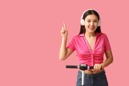 Beautiful young Asian woman in headphones with modern electric kick scooter pointing at something on...