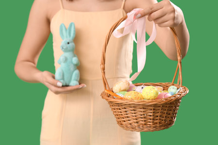 Young woman holding wicker basket with Easter eggs and toy rabbit on green backgroundの写真素材