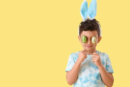 Cute little boy in bunny ears with Easter eggs on yellow background