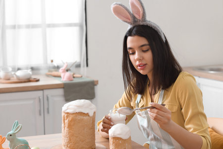 Beautiful young woman in bunny ears decorating Easter cakes in kitchenの写真素材