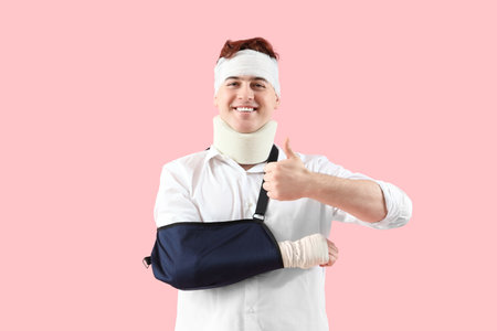 Injured young man after accident with broken arm showing thumb-up on pink backgroundの写真素材