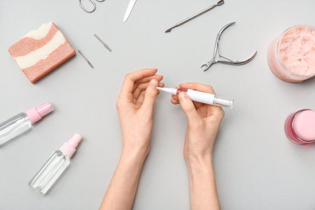 Female hands applying cuticle oil with pen, cosmetics and manicure instruments on gray backgroundの写真素材