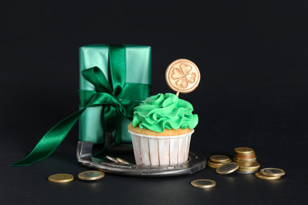 Tasty cupcake with gift box, horseshoe and golden coins for St. Patrick's Day on black backgroundの写真素材