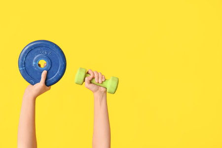 Female hands with barbell plate and dumbbell on yellow background, closeupの写真素材