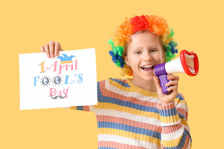 Funny little girl in clown wig with megaphone and card on yellow background. April Fools' Day celebrationの写真素材