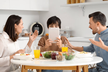 Teenage boy using tablet computer and his angry parents at table during dinner in kitchen. Family problem conceptの写真素材
